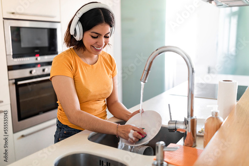 Smiling Arab Housewife Washing Dishes In Kitchen And Listening Music In Headphones