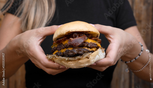 Monster burgers. Closeup view of a woman holding a hamburger with two meat medallions, cheddar cheese, bacon, grilled onions and cucumber.
