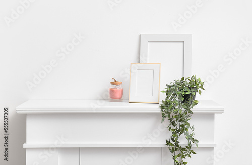 Blank photo frames, candle and houseplant on mantelpiece near white wall photo