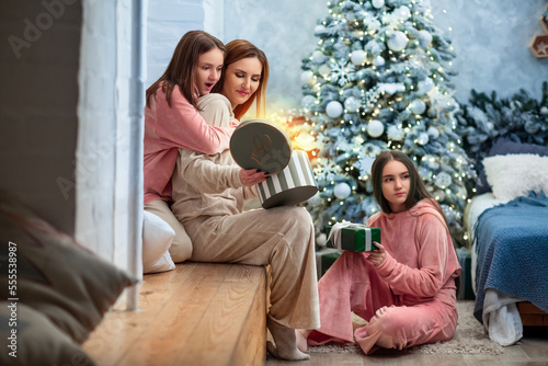 Young caucasian attractive brunette mother and her teenage pretty lovely daughters sitting on a windowsill in a cosy christmas room opening gift present box sincerely surprising