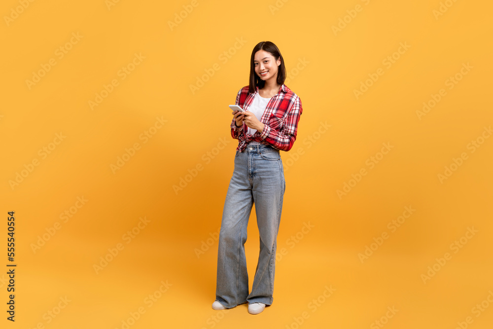 Full length shot of happy asian lady using smartphone and standing isolated over orange studio background, copy space Full length shot of happy asian lady using smartphone and standing isolated over orange studio background, copy space