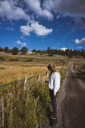Woman wearing a white sweater and black jeans picking flowers in front of a bright blue sky and yellow and green meadow in the fall - rural Rocky Mountain vibes