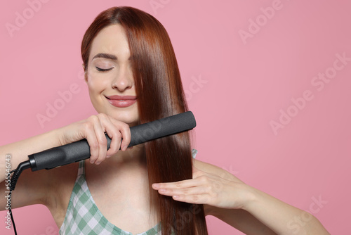 Beautiful woman using hair iron on pink background, space for text