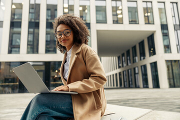 Smiling african business woman working laptop while sitting outside on background of office building