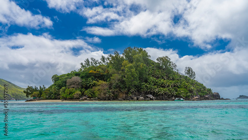 A secluded tropical island completely overgrown with tropical vegetation. Palm trees   picturesque boulders near the shore. A lonely boat is moored in the turquoise ocean. Blue sky  clouds. Seychelles