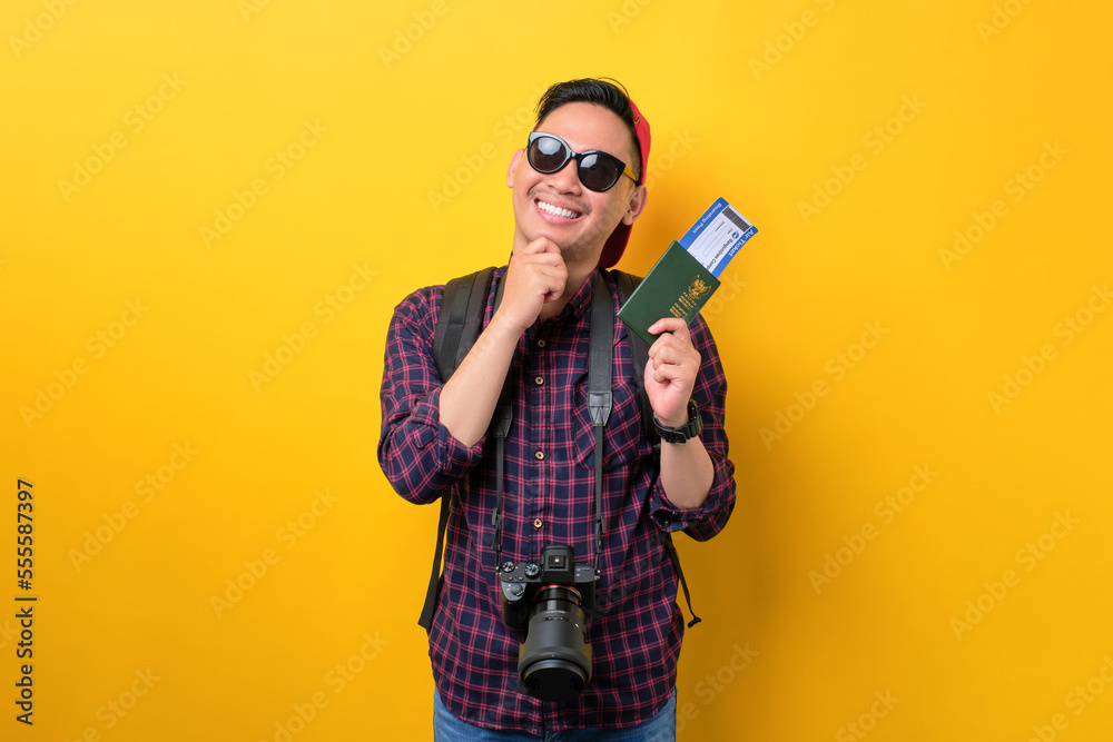 Thoughtful young Asian man with backpack holding passport and flight tickets, thinking of traveling isolated over yellow background. Tourism and holiday trip concept Thoughtful young Asian man with backpack holding passport and flight tickets, thinking of traveling isolated over yellow background. Tourism and holiday trip concept