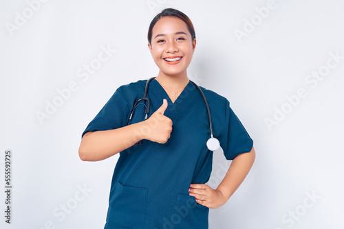 Smiling young Asian woman nurse wearing blue uniform with a stethoscope has all under control and shows thumbs-up in approval isolated on white background. Healthcare medicine concept