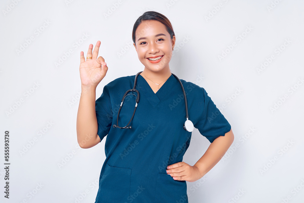 Smiling young Asian woman nurse wearing blue uniform with stethoscope, assures the quality of test results in their clinic lab, showing okay sign isolated on white background Smiling young Asian woman nurse wearing blue uniform with stethoscope, assures the quality of test results in their clinic lab, showing okay sign isolated on white background