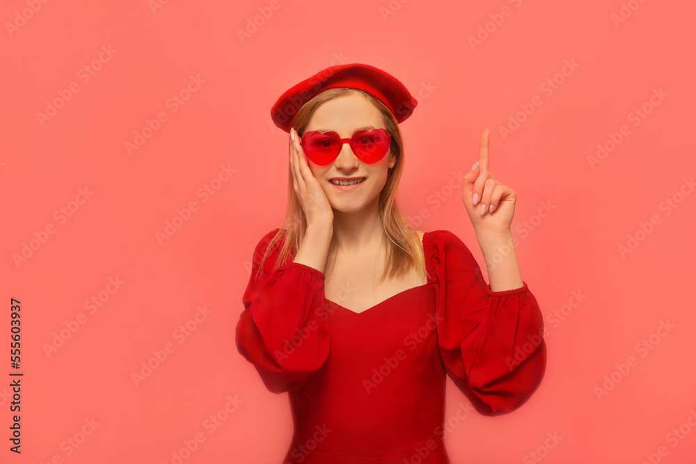 Happy smiling stylish girl pointing index finger up or holding finger up, keeping hand on face and looking at camera on pink background.
Young woman wearing red dress, beret and heart shaped glasses. Happy smiling stylish girl pointing index finger up or holding finger up, keeping hand on face and looking at camera on pink background.
Young woman wearing red dress, beret and heart shaped glasses.