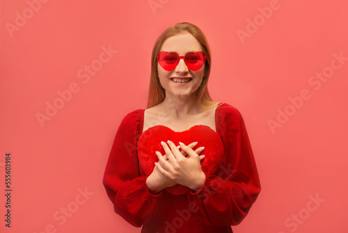 Excited happy smiling woman holding red toy heart close to chest, wearing red dress with trendy heart glasses and looking at camera isolated on pink background.

St Valentines Day celebration concept.