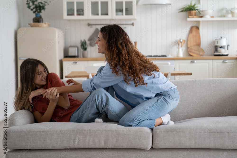 Cheerful Caucasian woman and teenage girl entertain themselves sits on sofa in spacious bright house. Friendly girlfriends different ages play tickling get sincere pleasure from joint rest and pastime Cheerful Caucasian woman and teenage girl entertain themselves sits on sofa in spacious bright house. Friendly girlfriends different ages play tickling get sincere pleasure from joint rest and pastime