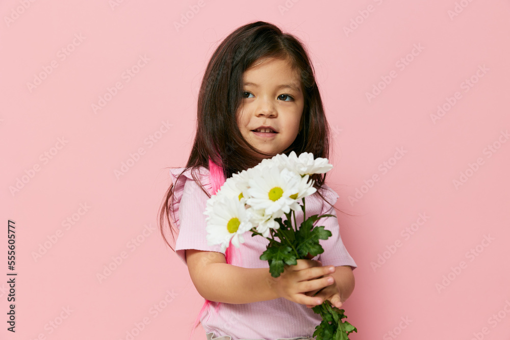 a little beautiful preschool girl stands on a pink background in a pink T-shirt smiling cheerfully holding a bouquet of white daisies in her hands standing sideways to the camera a little beautiful preschool girl stands on a pink background in a pink T-shirt smiling cheerfully holding a bouquet of white daisies in her hands standing sideways to the camera
