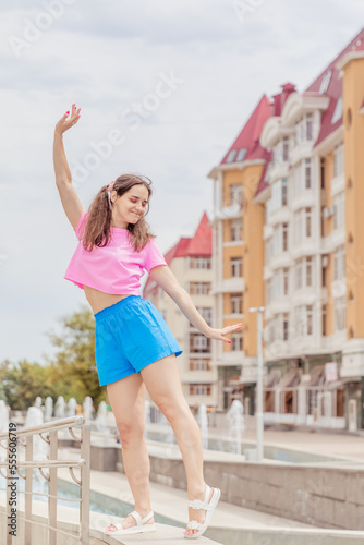 Beautiful girl in candy color clothes and bright makeup dancing and having fun near city buildings and fountain. Brunette girl posing on summer day during the walk in the center of Stavropol, Russia
