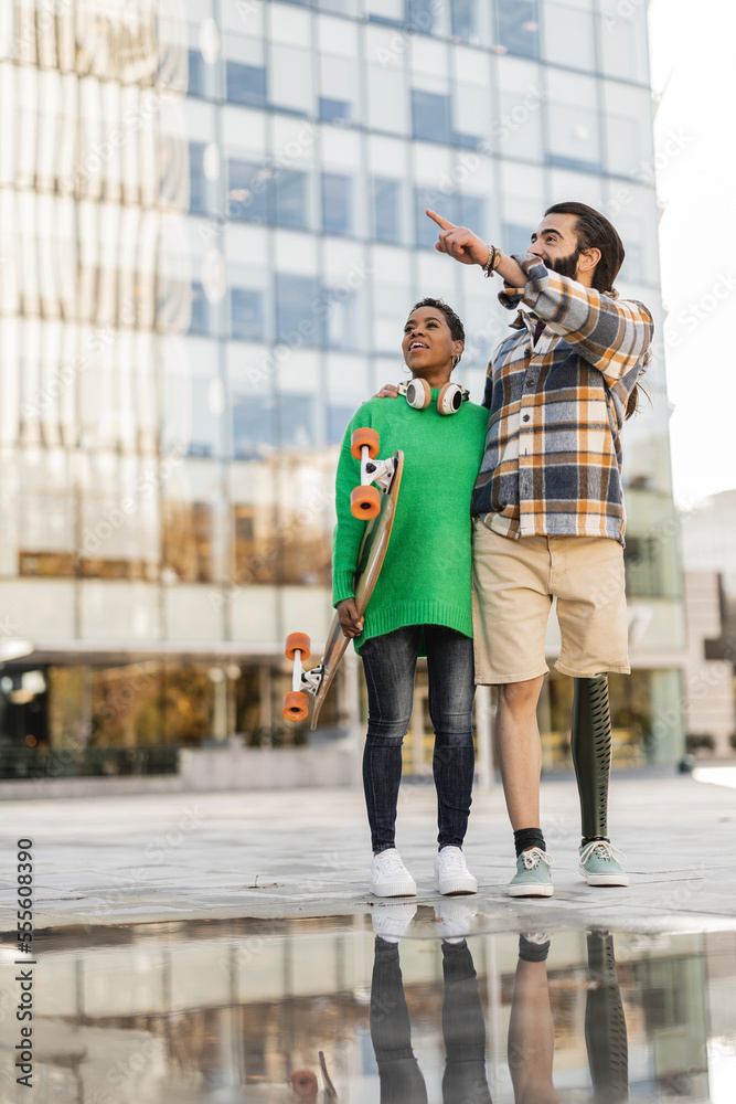Obraz premium funny couple with skateboard, in the city standing pointing, african american woman, hispanic man with amputated leg and a prosthesis.