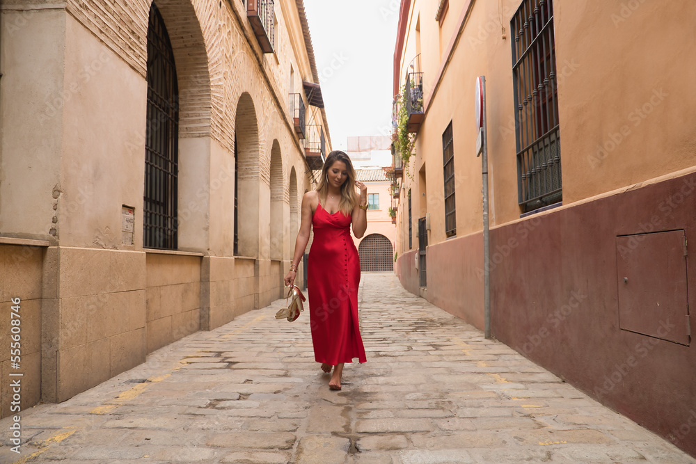 Young, attractive, blonde woman, wearing an elegant red party dress and holding golden high heels in her hand, walking barefoot down a city alley. Concept beauty, fashion, elegance, luxury.