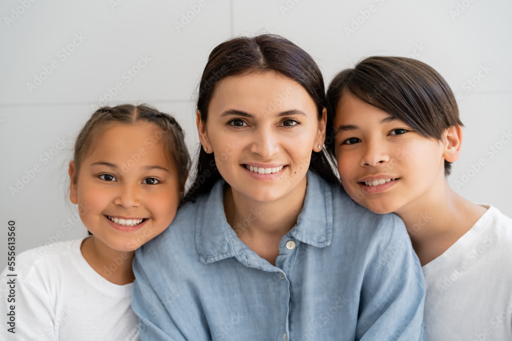 Positive woman and asian children looking at camera near wall at home Positive woman and asian children looking at camera near wall at home