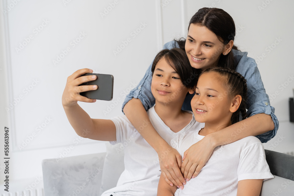 Smiling mother hugging asian kids while son taking selfie at home Smiling mother hugging asian kids while son taking selfie at home