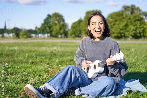 Portrait of beautiful asian woman singing, playing ukulele guitar in park, sitting alone on grass on sunny day photo