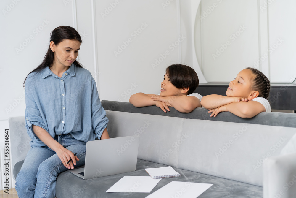 Smiling asian kids looking at mother using laptop near documents at home Smiling asian kids looking at mother using laptop near documents at home