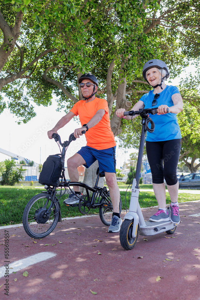 Fototapeta premium coppia di anziani coniugi vestiti sportivi e con caschetto di protezione in testa si muovo con mezzi elettrici nella pista ciclabile
