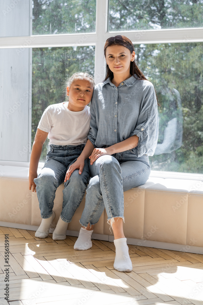 Smiling woman looking at camera near asian daughter and window at home