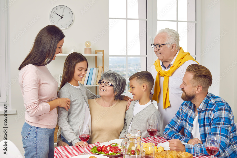Cheerful family gather all together for a cozy dinner meal at home. Happy multi generational family meet and have fun together. Grandmother hugging her little grandchildren who visit her with parents