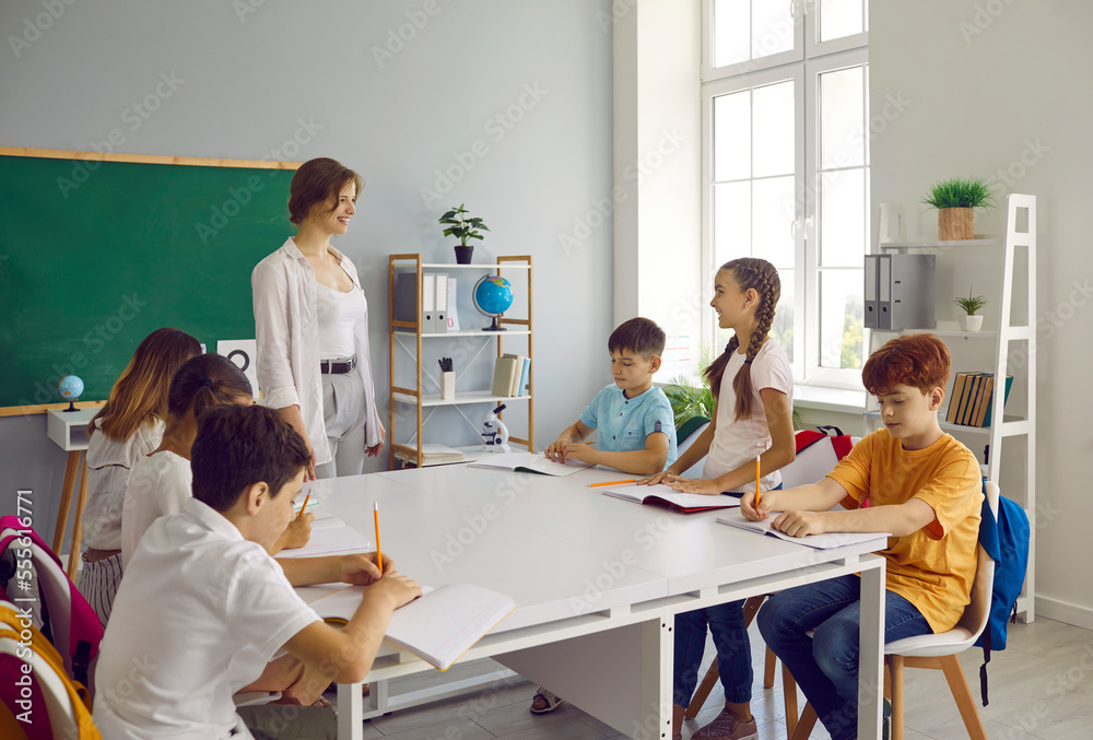Obraz premium Elementary school students do their homework together in extended day group class. Small schoolgirl answers questions of her friendly female teacher surrounded by children sitting at one large table.