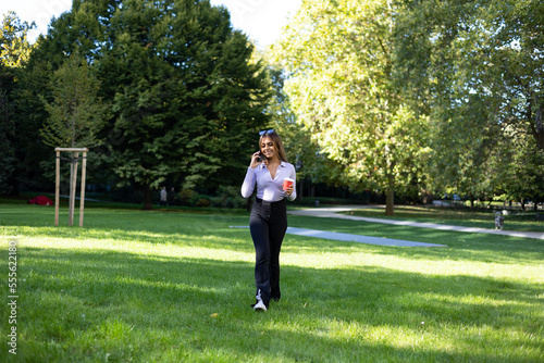 Woman talking on phone with smartphone. Portrait woman with brown hair, cup of coffee in hand and cell phone. Outdoor, public park, relaxed position and summer. 