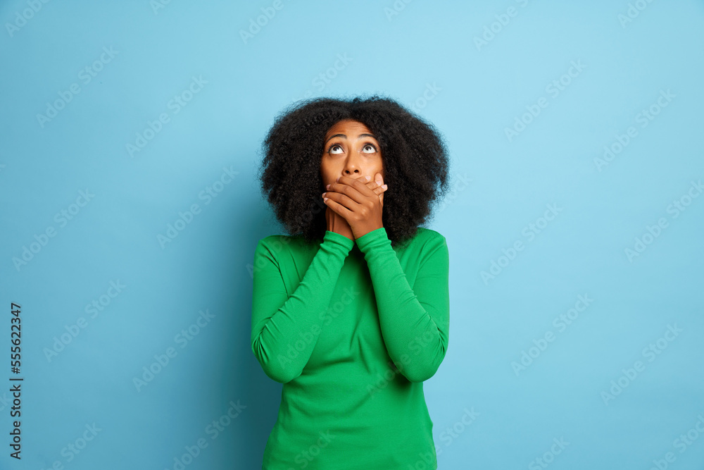 Woman in green jumper, closed her mouth, she can not talk much about, looks up, scared to say the truth, isolated next to blue wall