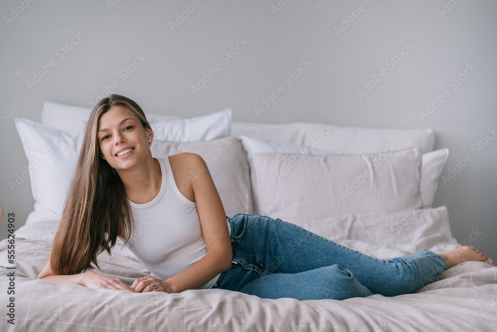 Attractive Spanish young adult woman in white t-shirt and blue jeans laying on bed with big cushions looks at camera toothy smiling. Mockup, cozy home. Successful lady relaxing at home. Attractive Spanish young adult woman in white t-shirt and blue jeans laying on bed with big cushions looks at camera toothy smiling. Mockup, cozy home. Successful lady relaxing at home.