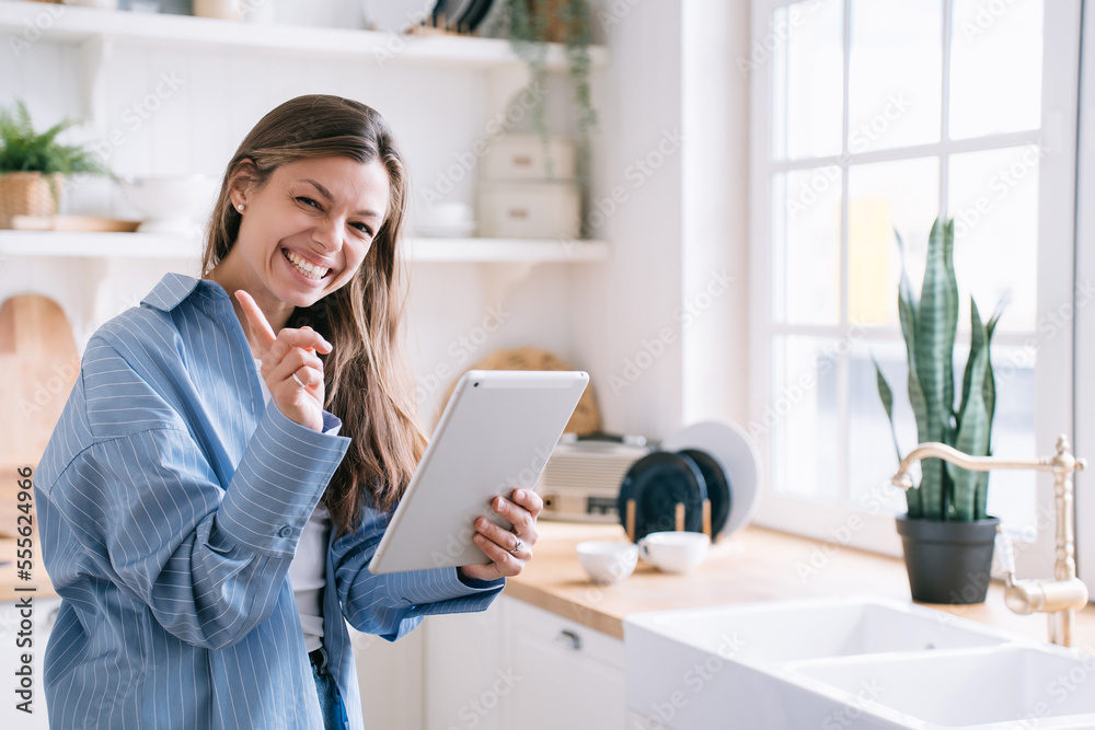 Cheerful young adult caucasian woman in blue shirt holds tablet toothy smiles looks at camera warns by index finger not to look at her messages, tablet screen. Secret concept. Girl smiles at kitchen. Cheerful young adult caucasian woman in blue shirt holds tablet toothy smiles looks at camera warns by index finger not to look at her messages, tablet screen. Secret concept. Girl smiles at kitchen.