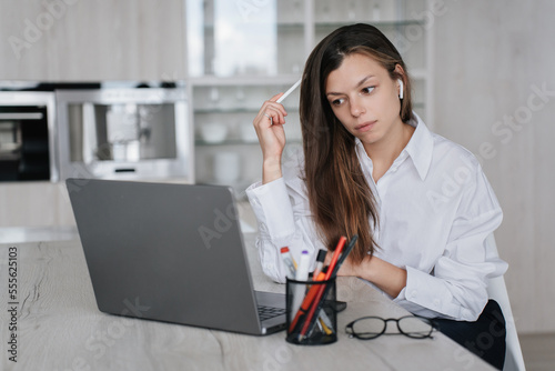 Perplexed young adult woman white shirt work at home using laptop thinking about new project looks at screen tries to resolve problem. Overloaded student girl remote learning. Business and education.