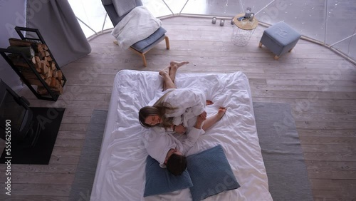 Top view joyful young Caucasian couple having fun cuddling on bed indoors. Wide shot carefree relaxed boyfriend and girlfriend in bathrobes laughing tickling in bedroom at home