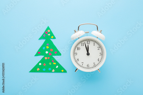 White alarm clock and green Christmas tree with colorful balls created from paper on light blue table background. Pastel color. Countdown to midnight before New Year. Closeup. Top down view.