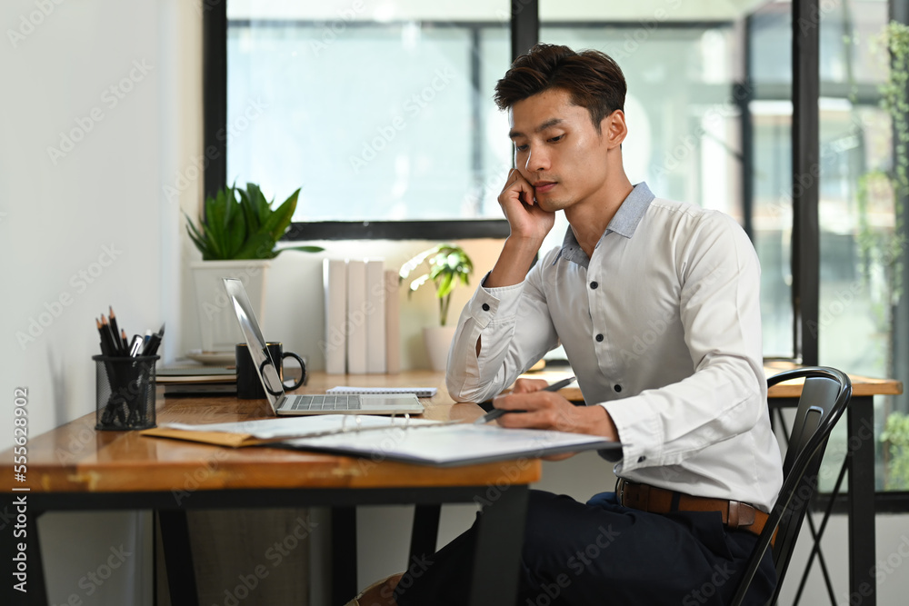 Bored asian businessman working with financial reports and using laptop at office desk Bored asian businessman working with financial reports and using laptop at office desk