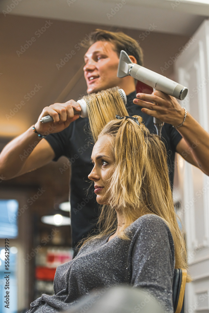 Fototapeta premium Hairstylist appointment. Young blond woman having her hair blown dried by professional hairdresser. Focus on the foreground. Vertical indoor portrait. Copy space. High quality photo