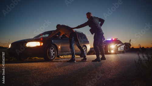 A Professional Middle Aged Policeman Performing a Pat-Down Search on a Fellon With his Hands on Car Hood. Documentary-like Shot of Procedure of Arresting Suspects. Experienced Cop Looking for Weapons