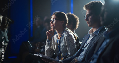 Young Woman Sitting in a Crowded Audience at a Business Conference. Female Attendee Writing Down Important Bullet Points into a Notebook. Keynote Speech in Auditorium with Successful Businesspeople. photo