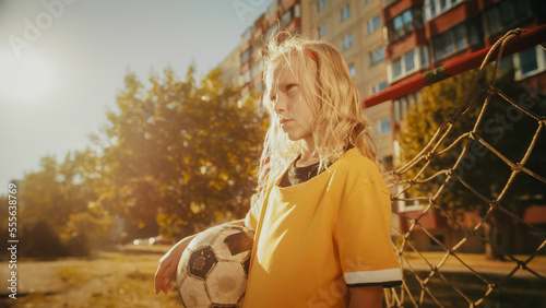 Portrait of a Serious Blond Girl in Yellow T-Shirt Holding a Soccer Ball in the Neighborhood, Standing Next to a Goal Gate. Young Football Player Looking at Camera. Kids Playing in the Background. © Gorodenkoff