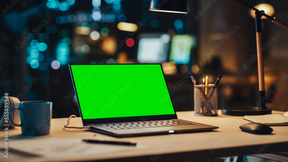 Laptop Standing on a Desk with a Green Screen Chromakey Mock Up Display. Table with Computer, Coffee Mug, Headphones and Notebook in Creative Office at Night.