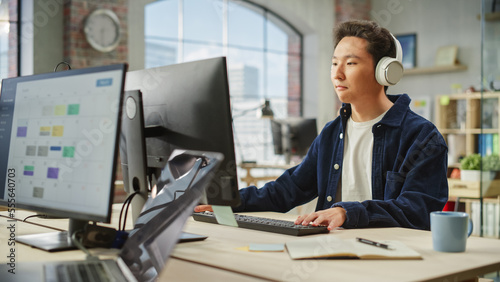 Portrait of Asian Male Employee Working on Computer in a Modern Office During the Day. Creative Graphic Designer Inspired While Listening to Music on Headphones.