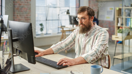 Portrait of Young Caucasian Bearded Man with Glasses Focused While Sitting and Working on Computer in a Bright Spacious Office. Male Legal Consultant Typing Notes on Keyboard.