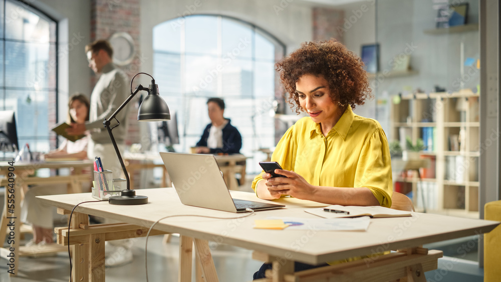 Portrait of Young Biracial Woman Working With a Laptop in a Bright Spacious Office. Female Production Lead Receiving an Important Work Notification on her Smartphone. Portrait of Young Biracial Woman Working With a Laptop in a Bright Spacious Office. Female Production Lead Receiving an Important Work Notification on her Smartphone.