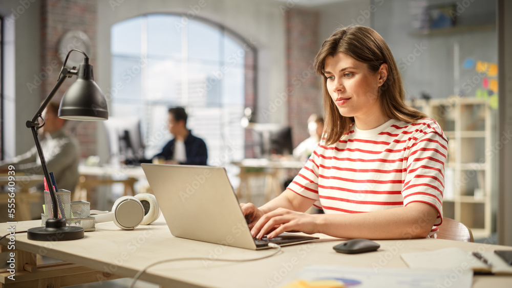 Portrait of Caucasian Young Inspired Woman Smiling and Working on Laptop in Busy Office. Analyst Gathering Data and Being Productive. People Working on Computers in the Background. Portrait of Caucasian Young Inspired Woman Smiling and Working on Laptop in Busy Office. Analyst Gathering Data and Being Productive. People Working on Computers in the Background.
