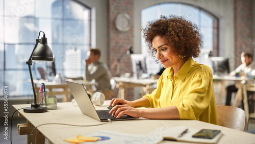 Portrait of Focused Young Woman Smiling and Using Laptop in a Dynamic Modern Office. Female Accountant Developing a Strategic Plan. Colleagues Working on Computers in the Background.