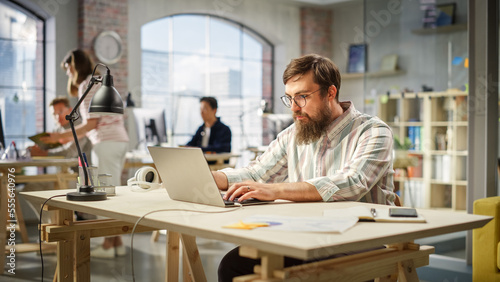 Portrait of Motivated Caucasian Young Man With Beard Working on a Laptop in Office. Male Director Preparing a Marketing Project. Diverse Team Working on Computers in the Background.