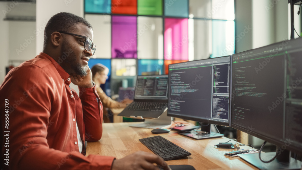 Black Man Writing Lines of Code On Desktop PC With Two Monitors and a Laptop in Stylish Office. Professional Male Developer Programming Artificial Intelligence Software for Start-Up Company.
