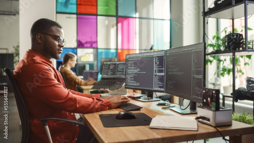 Black Man Writing Lines of Code On Desktop PC With Two Monitors and a Laptop Aside in Stylish Office. Professional Male Developer Programming Artificial Intelligence Software for Start-Up Company.