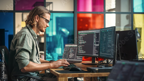 Obraz na płótnie Male Programmer Writes Code On Desktop Computer With Multiple Monitor Setup And a Laptop Aside In Creative Office