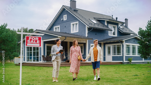 Excited Young Couple Chatting with Happy Real Estate Agent. Businesswoman Talking About the House with Young Man and Pregnant Female. Standing on a Lawn Next to For Sale Sign on a Warm Day.